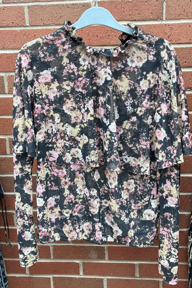 Floral-patterned long-sleeve blouse by Ayanapa on a hanger against a brick wall.
